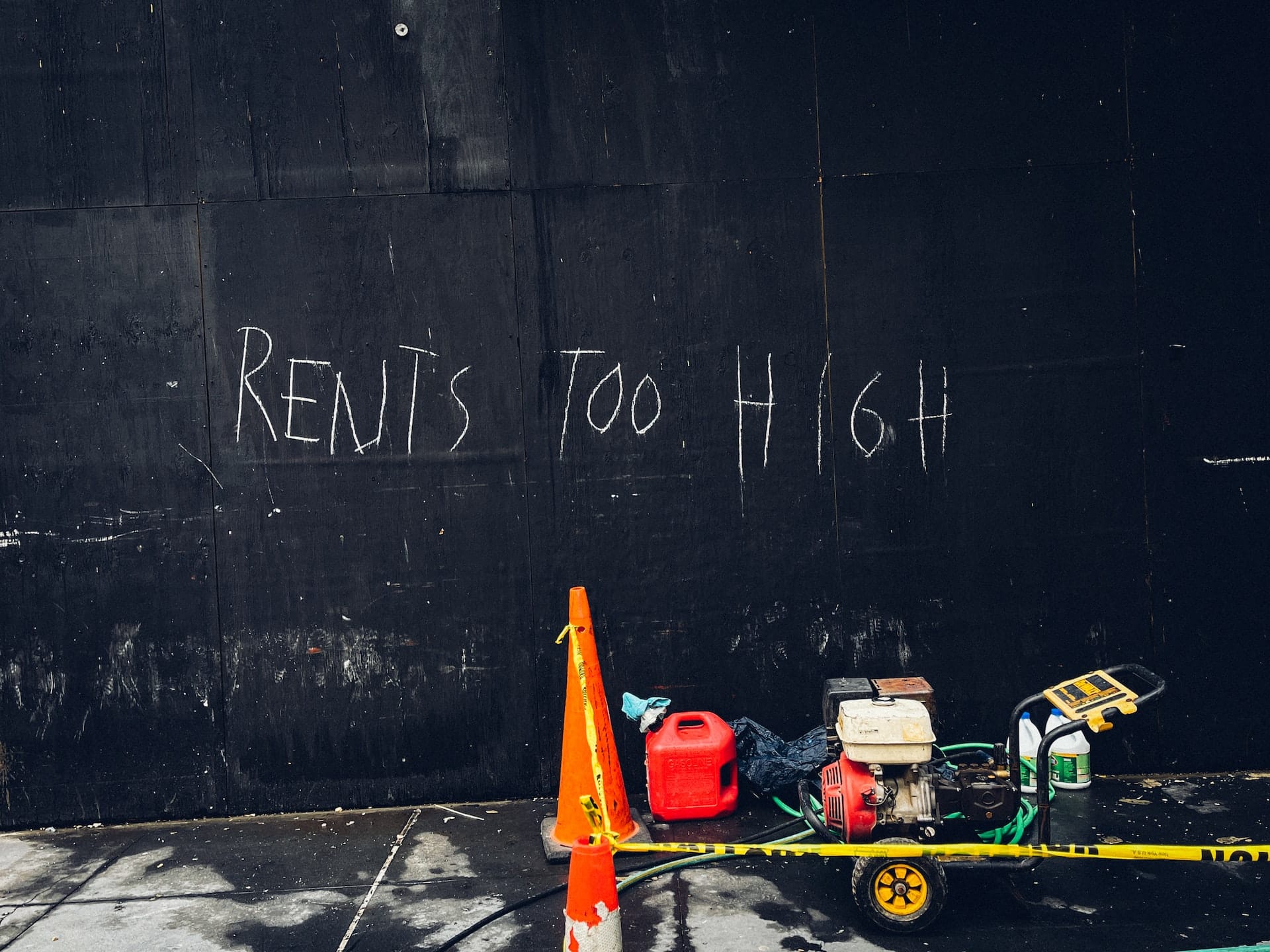 Black wall with the words "rents too high" written on it. An orange cone, red gas can, yellow caution tape, and generator in front of black wall.