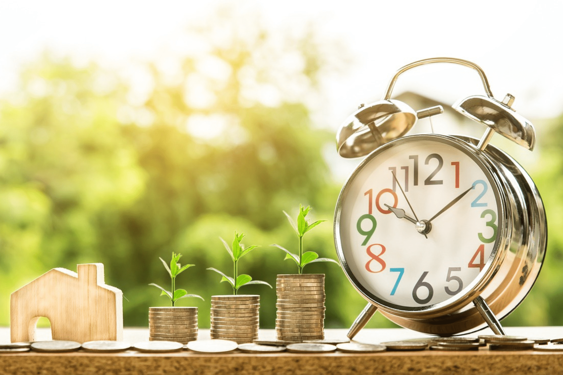 A close-up of an alarm clock, a stack of coins, and a miniature house sitting atop a wooden table. Green leaves are growing from the stack of coins. This symbolizes the financial benefits of Austin real estate investing.