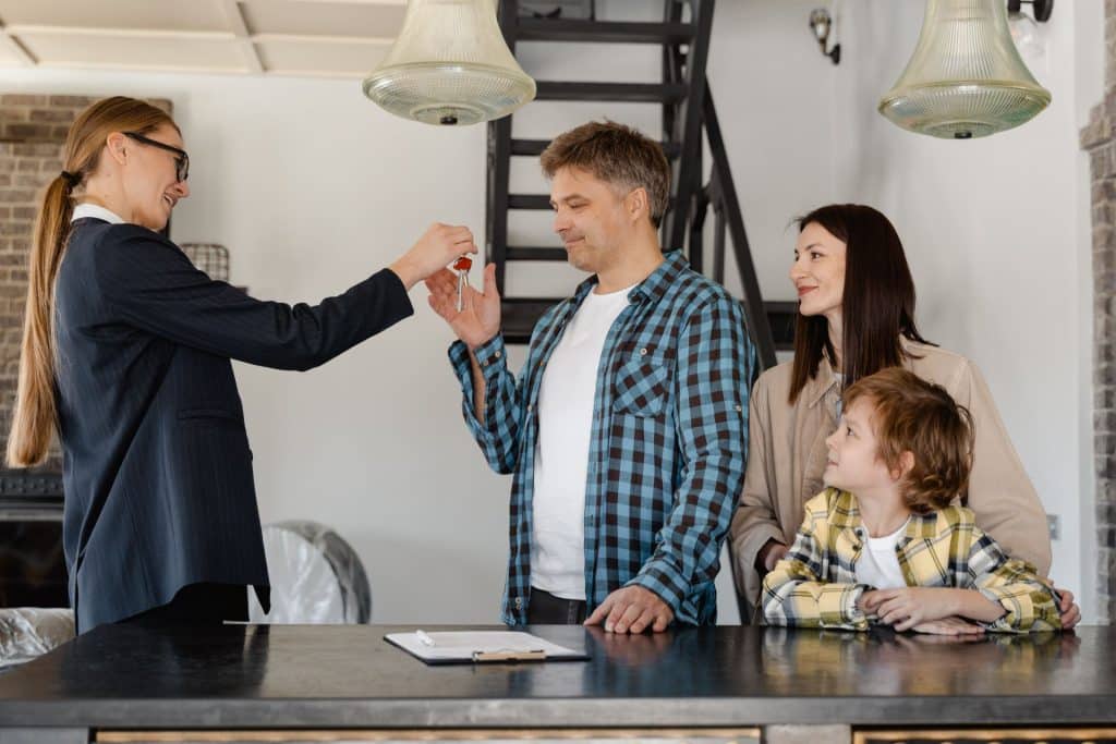 A property owner who is managing tenants solo hands over the property keys to the tenant father wjo is standing on the left side of his wife and daughter