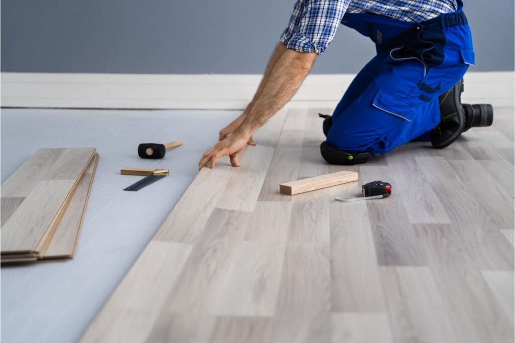 Property maintenance contractor installing light-colored laminate flooring as a flooring investment, kneeling in blue overalls with tools and stacked planks nearby.