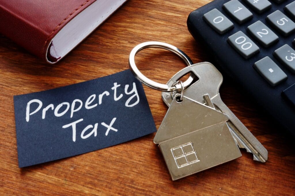 A black card with "Property Tax" written in chalk sits next to a house keychain, a calculator, and a book, illustrating a property tax protest.