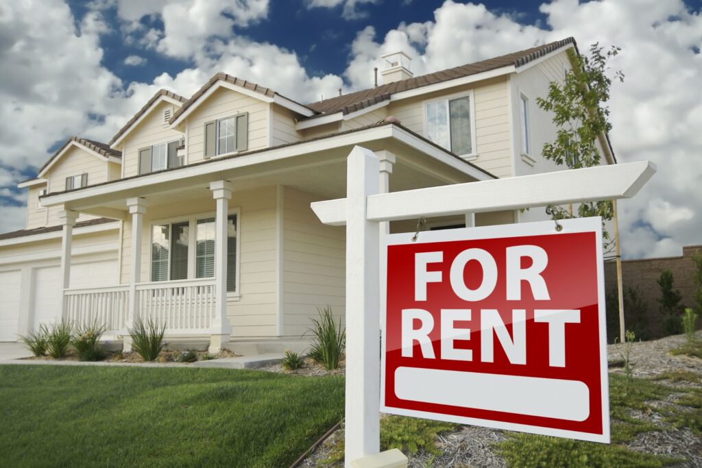 A two-story light yellow house with white trim and gray roof, ideal for a first time landlord. It has a well-kept lawn and landscaping. A red "For Rent" sign is in the front yard. The sky is blue with white clouds.