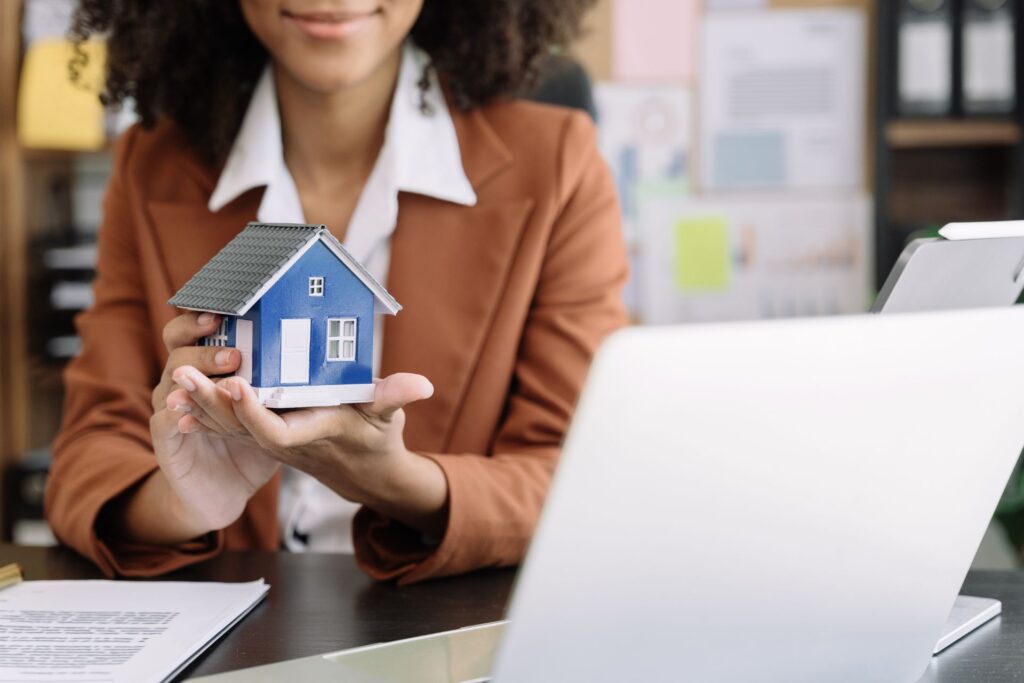 A woman holding a small, blue model house in her hands, symbolizing a rental walkthrough. She wears a brown blazer and white shirt, with a laptop and office background.