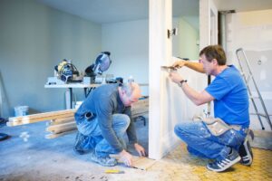 Two men working indoors on a rental property renovation. One is crouching, laying flooring, while the other kneels measuring or working on a wall or door frame. Tools, materials, a ladder, and an unfinished wall are visible, indicating ongoing construction.