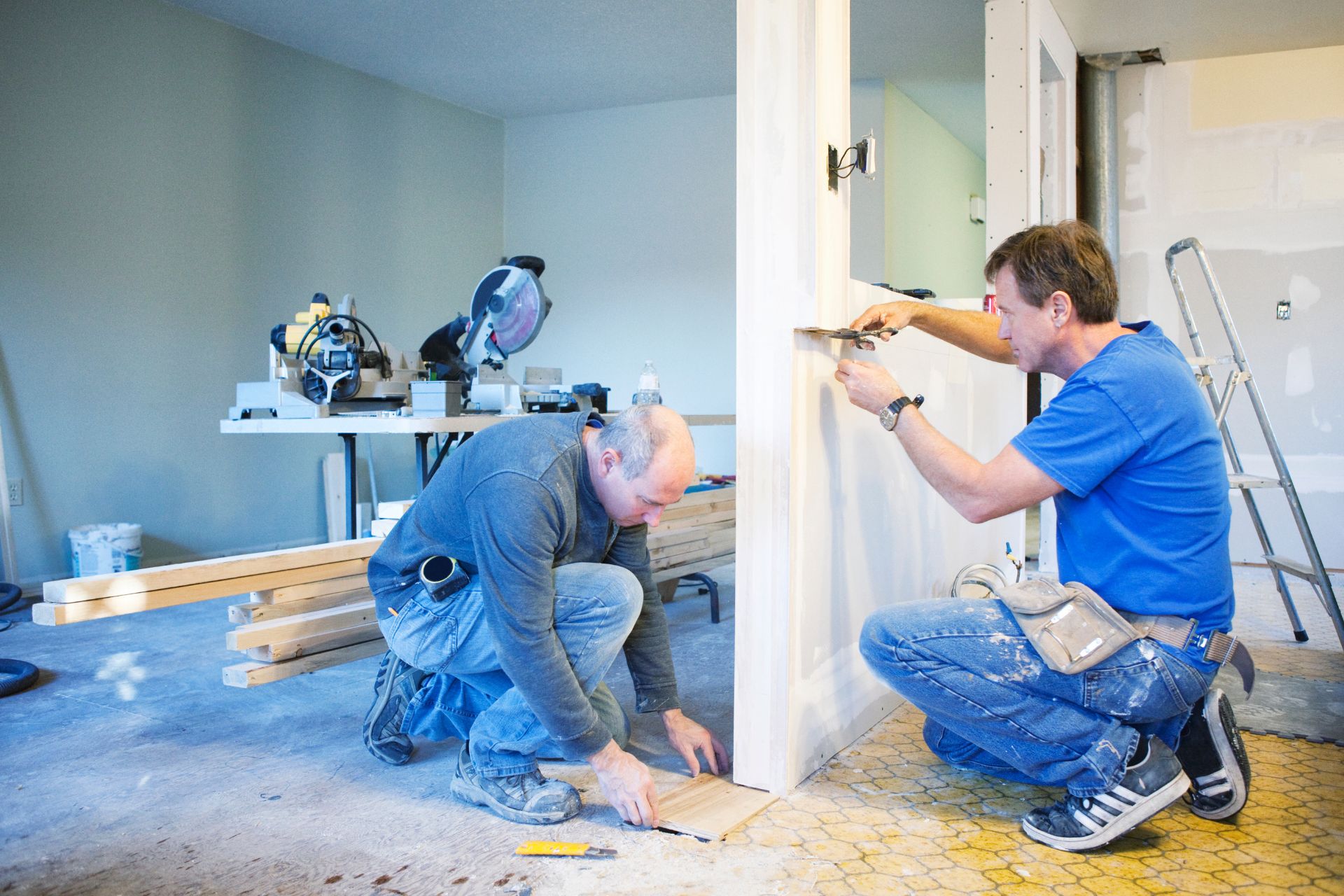 Two men working indoors on a rental property renovation. One is crouching, laying flooring, while the other kneels measuring or working on a wall or door frame. Tools, materials, a ladder, and an unfinished wall are visible, indicating ongoing construction.